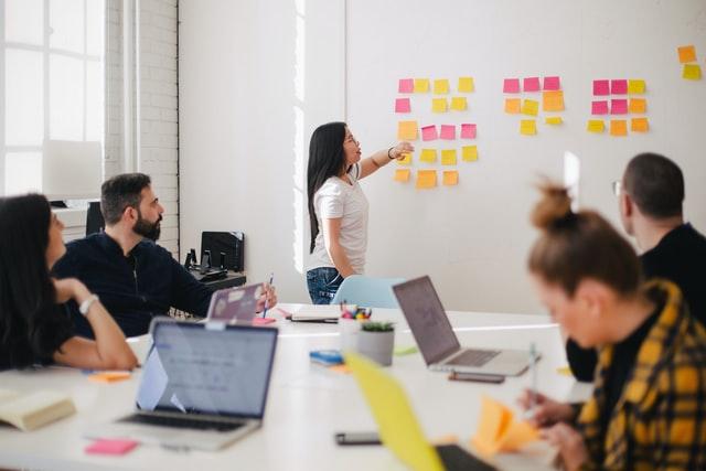 woman placing sticky note on whiteboard in front of colleague to discuss idea in meeting