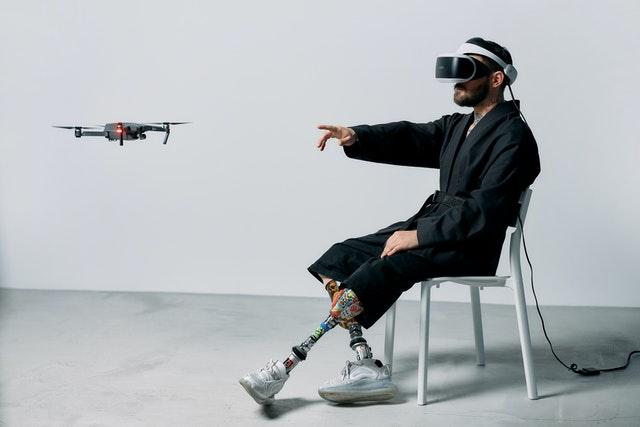 man with prosthetic leg sitting on a chair with virtual reality gear on face pointing a drone